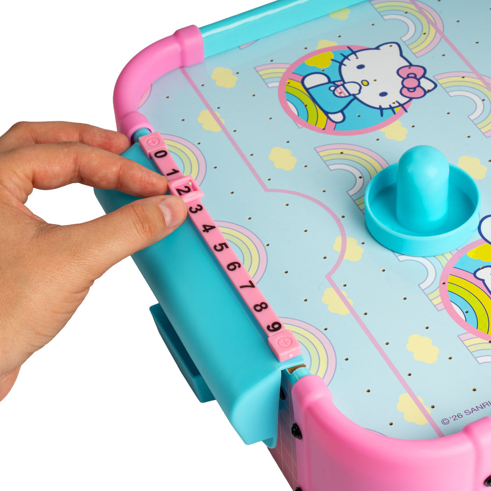 A close-up of a Hello Kitty toy game board with a hand adjusting pink numbered tiles on a turquoise and pink frame. The board features Hello Kitty illustrations, rainbows, and a turquoise spinner.