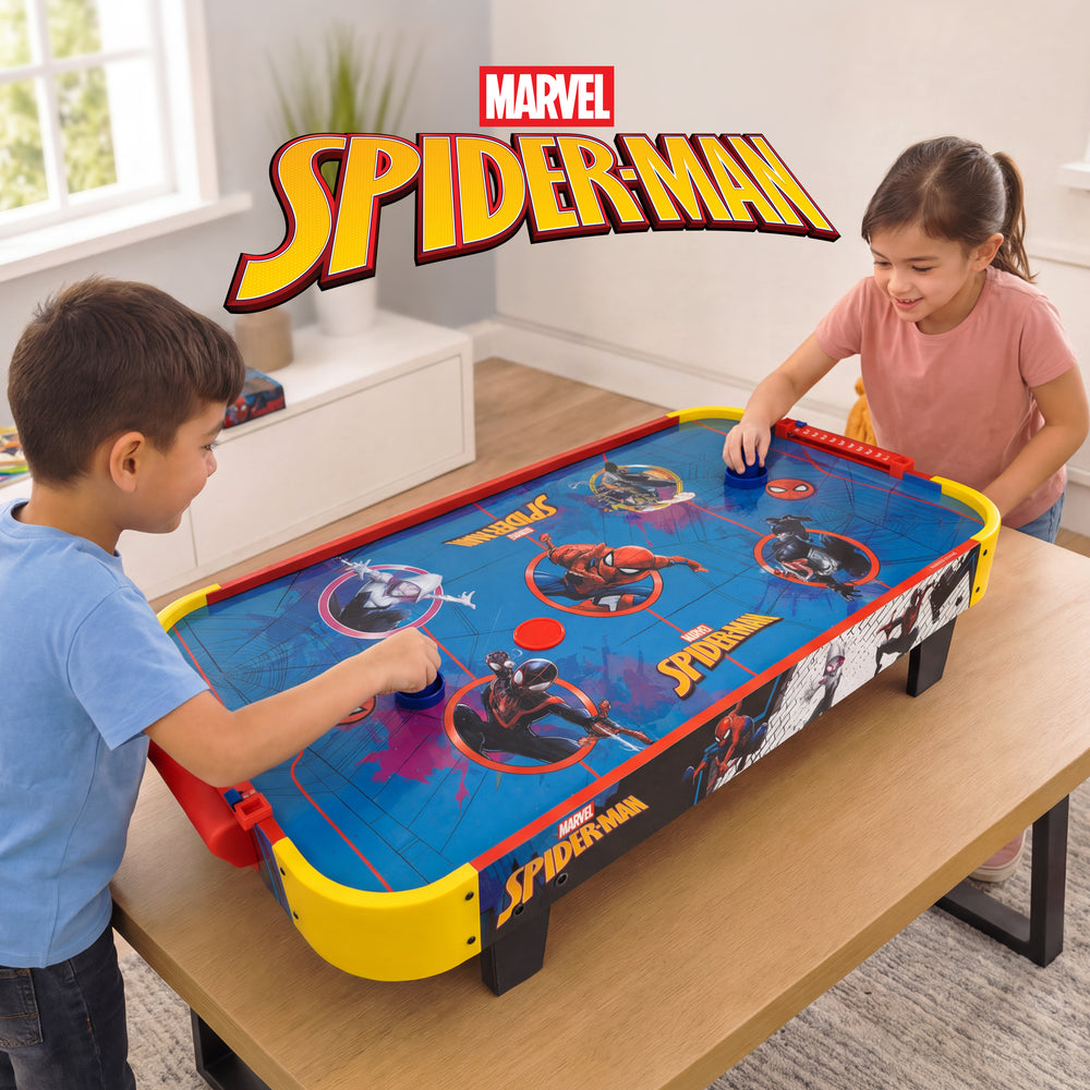 A Marvel Spider-Man air hockey table game shown in a child's room with two kids playing. The product is a rectangular table with a blue playfield featuring Spider-Man graphics, yellow and red borders, and black legs.