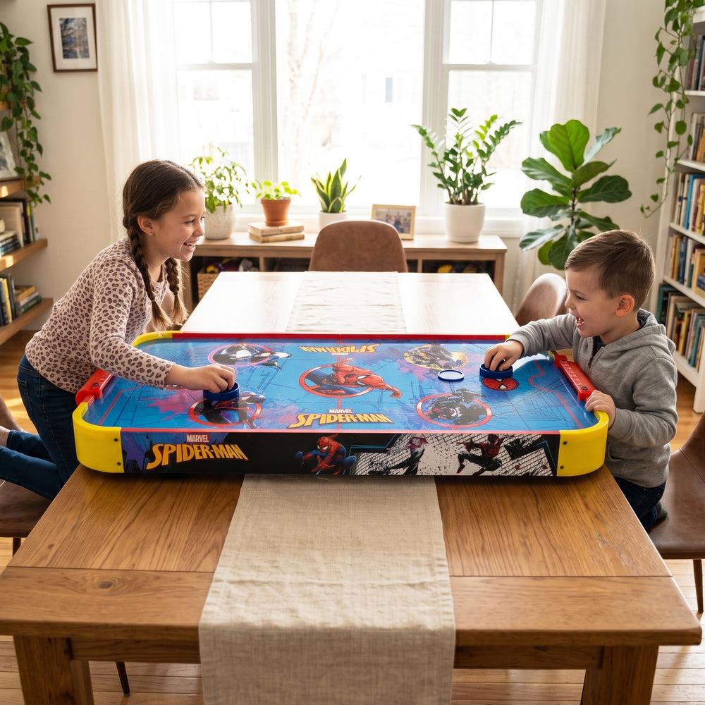 A children's Spider-Man air hockey game on a wooden dining table, with a girl and a boy playing in a bright, plant-filled room.