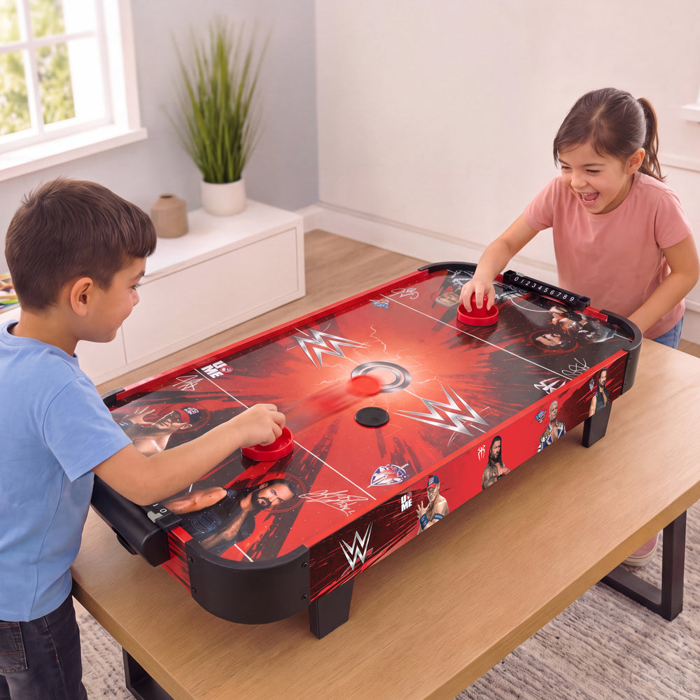 Two children playing an indoor WWE-themed air hockey game on a wooden table in a bright room. The red and black game board features WWE logos and player graphics, with the children holding red discs.