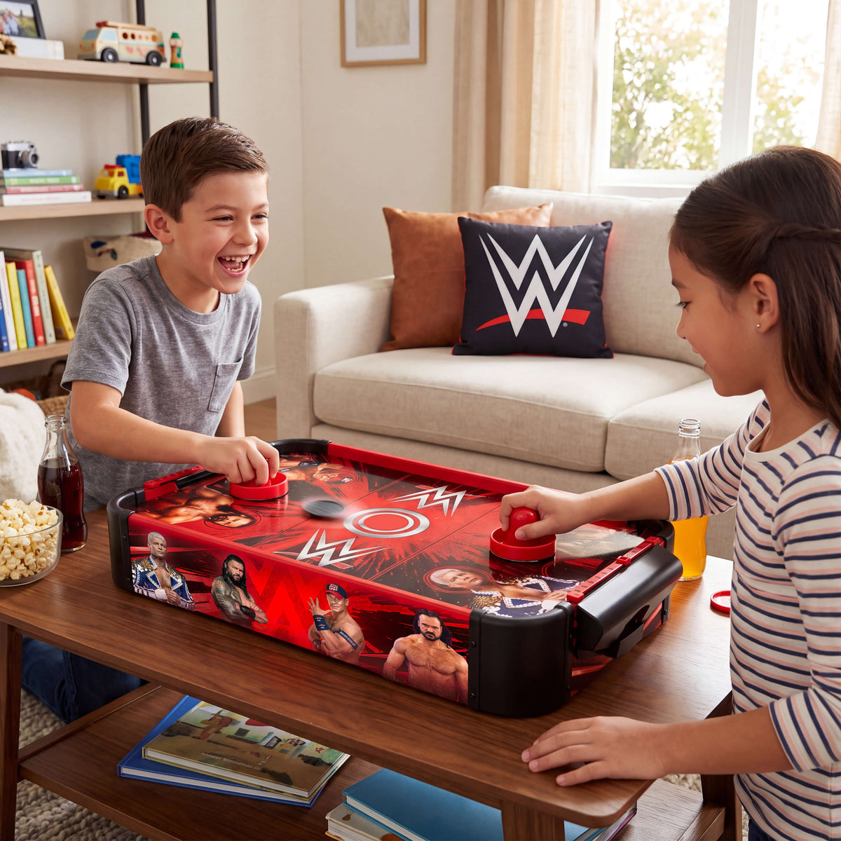A boy and a girl play WWE-themed air hockey on a red table in a cozy living room setting. The table features various WWE wrestler graphics, and a WWE pillow rests on the sofa in the background.