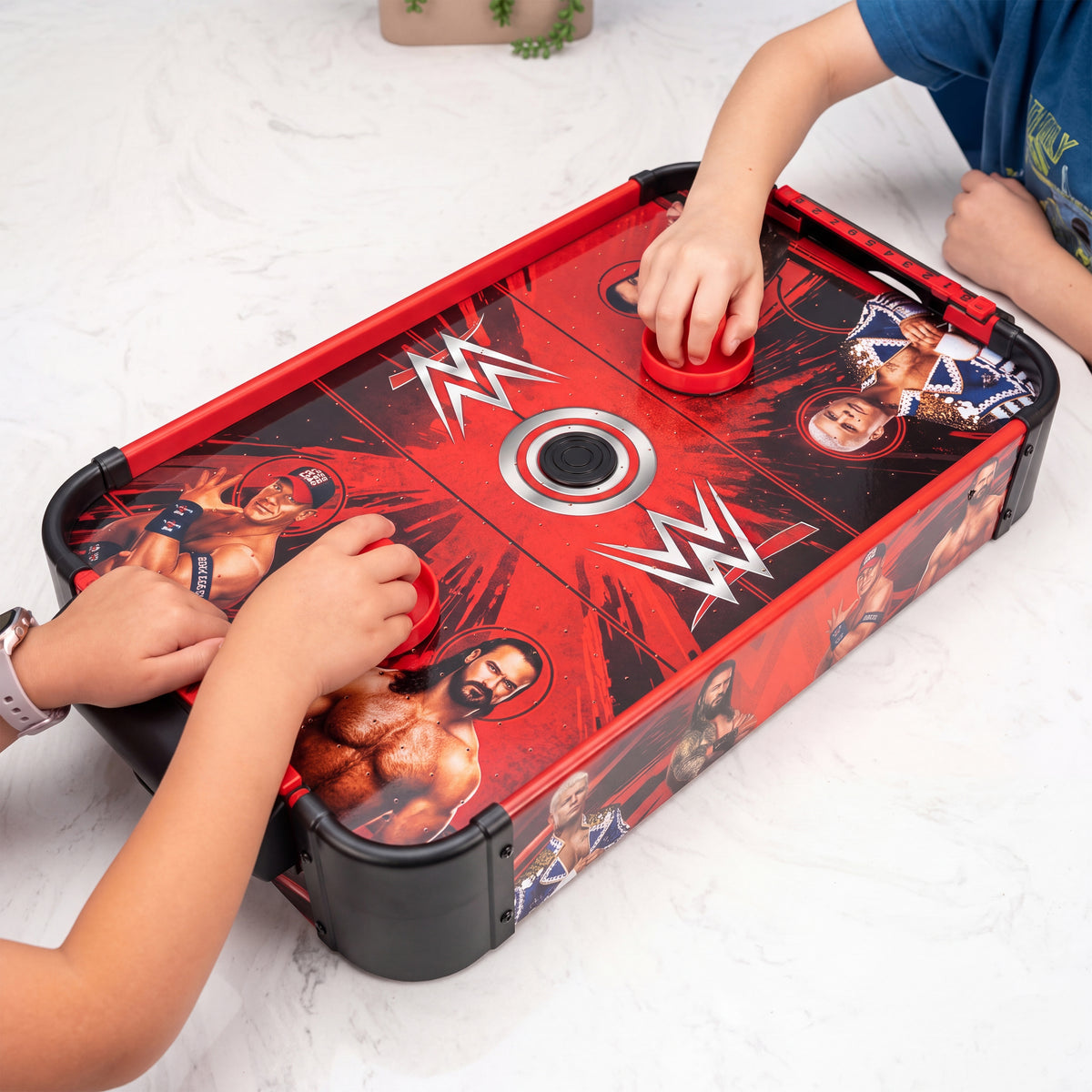 A WWE-themed air hockey table being played by two children on a white marble floor. The table features red borders, black corner bumpers, and a red and black graphic design with white WWE logos and wrestler portraits.