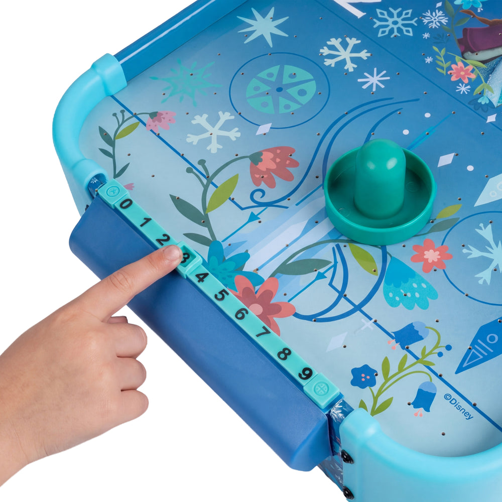 A close-up of a children's ice game board with a blue and turquoise design, floral and snowflake graphics, a green puck, and a finger pointing to the measurement strip along the side. A Disney logo is visible on the board.
