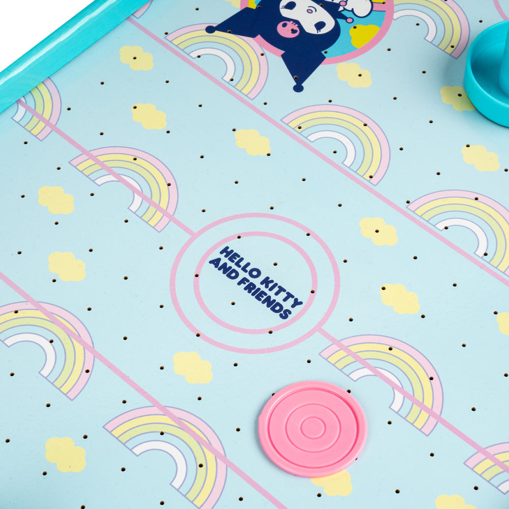 Close-up of a Hello Kitty and Friends themed game board or playmat with pastel rainbow patterns, yellow clouds, black dots, and a pink circular token. The text 'HELLO KITTY AND FRIENDS' is printed in the center, and a turquoise frame is visible around the edge.