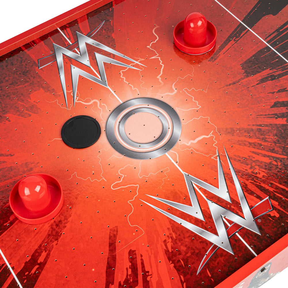 A close-up overhead view of a red WWE-themed air hockey table, showing a lightning-bolt style WWE logo, a central metal ring, a black puck, and red pucks on a red graphic surface.